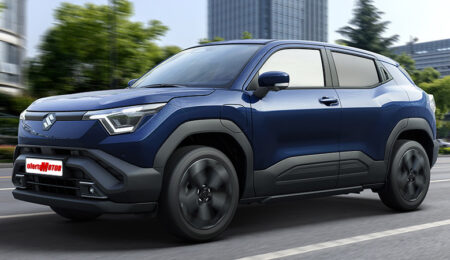 Un SUV Suzuki e Vitara azul del año 2025 está estacionado en una carretera con edificios y árboles al fondo.